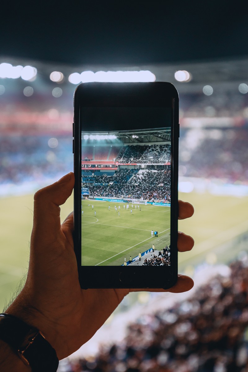 Man erkennt einen Mann, der ein Foto von dem laufenden Fußballspiel mit seinem Handy aufnimmt